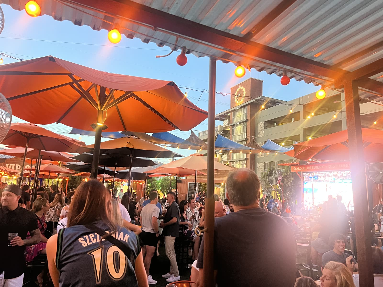 Crowded patio at OHSO Brewery in downtown Gilbert at sunset, with orange umbrellas and a big screen